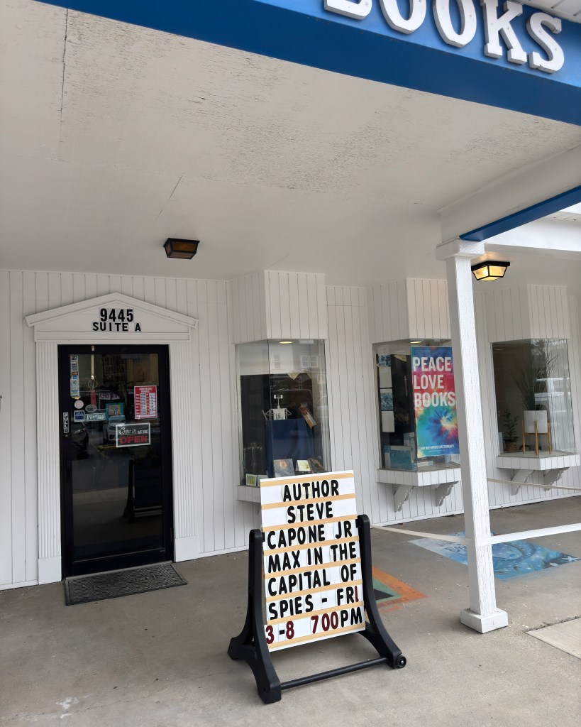 pictured is the front of a bookstore with its "open" or "closed" sign and a portable marquis with the words "author steve capone jr max in the capital of spies - Fri 3/8 7:00pm".