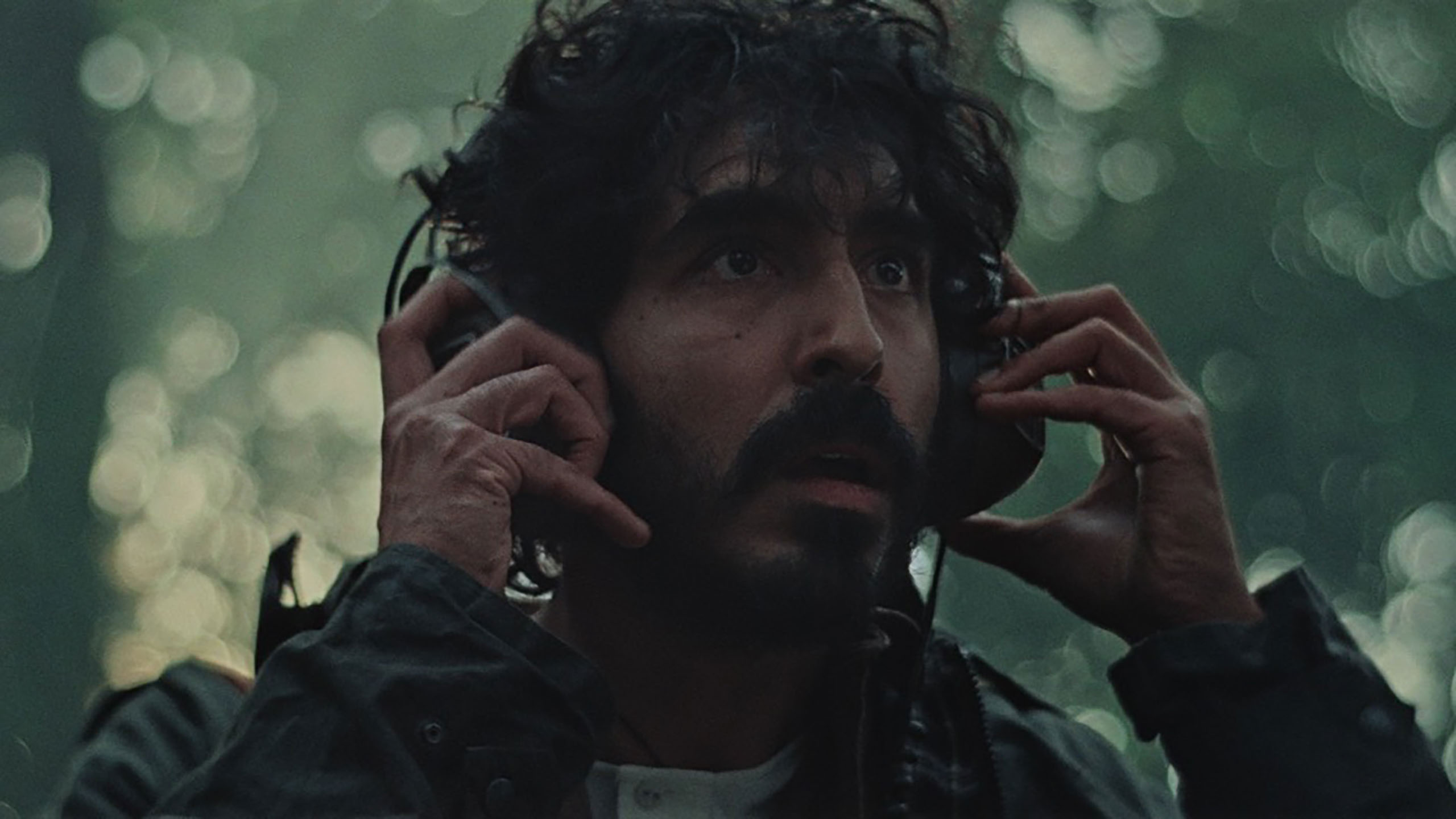 a nervous looking man with scruffy hair and a beard holds both hands on his over-the-ear headphones while looking around in a forest at something we cannot see